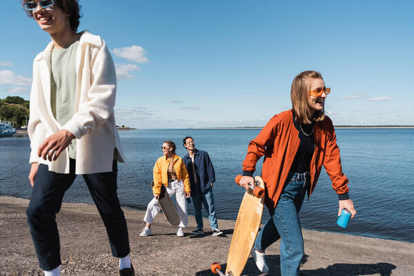 smiling woman walking on riverside with longboard and soda can near interracial friends