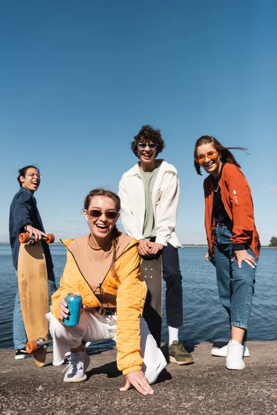 cheerful woman with soda can looking at camera near trendy friends on lake shore