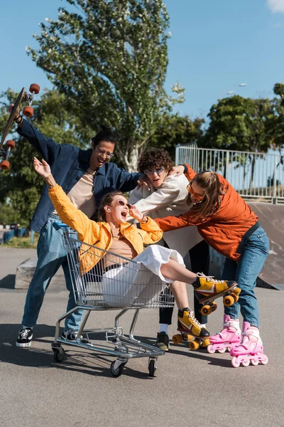 異人種間の友人の近くのショッピングカートに乗っている間に叫んでローラースケートで驚きの女性 — ストック写真