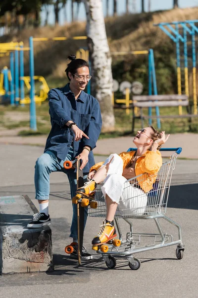 Aziatische Man Met Longboard Wijzend Naar Lachende Vrouw Zittend Winkelwagen — Stockfoto