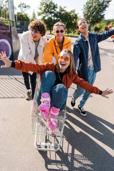 Thrilled Woman Outstretched Hands Riding Shopping Trolley Interracial Friends — Stock Photo, Image