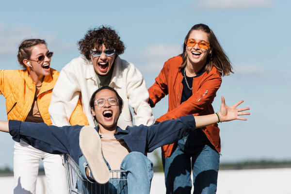 thrilled asian man with outstretched hands shouting in shopping cart near friends