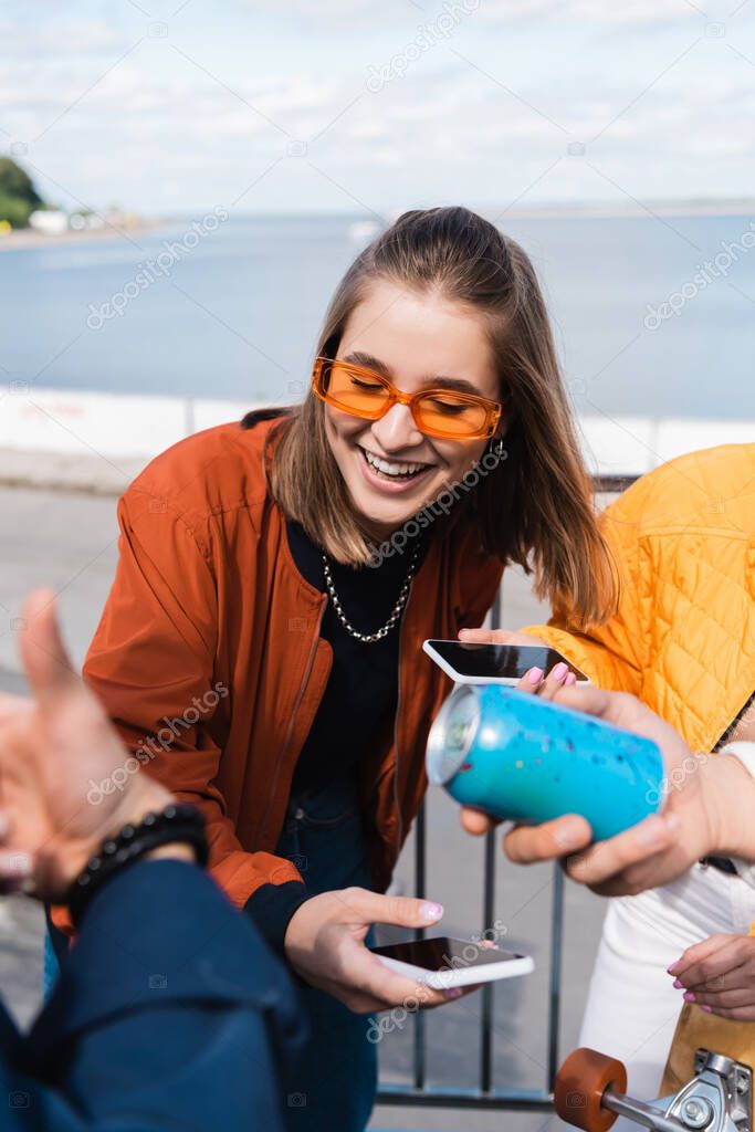 smiling woman in trendy sunglasses holding smartphone with blank screen near friends