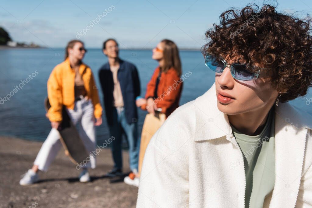 young man in trendy eyeglasses looking away near friends on blurred background