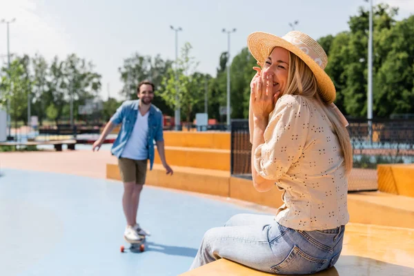 陽気女性で太陽帽子座っています近くにぼやけたボーイフレンドライディングLongboardでスケートパーク — ストック写真