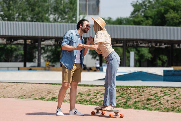 Smiling man in sunglasses holding hands of girlfriend rising longboard in park 