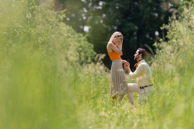 Side view of stylish man holding hand of girlfriend while kneeling in summer park  clipart