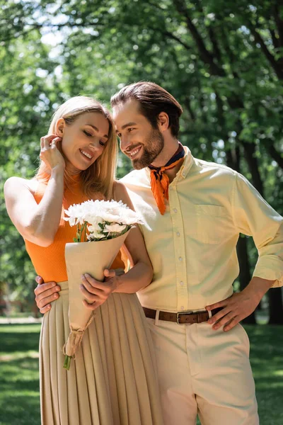 Sorrindo Homem Abraçando Namorada Elegante Com Flores Parque — Fotografia de Stock