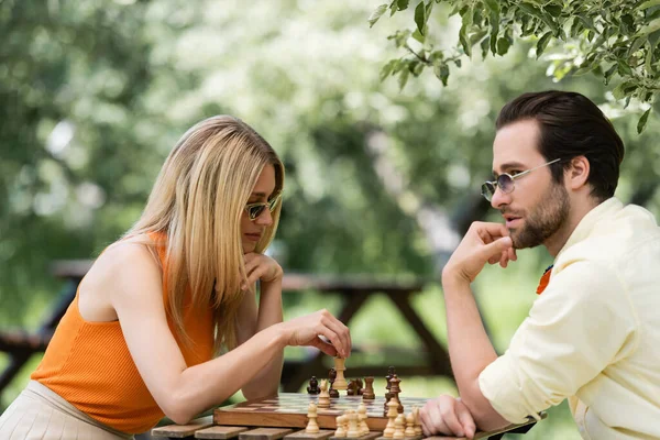 戴着太阳镜的时髦女人在公园和年轻的男朋友下棋 — 图库照片