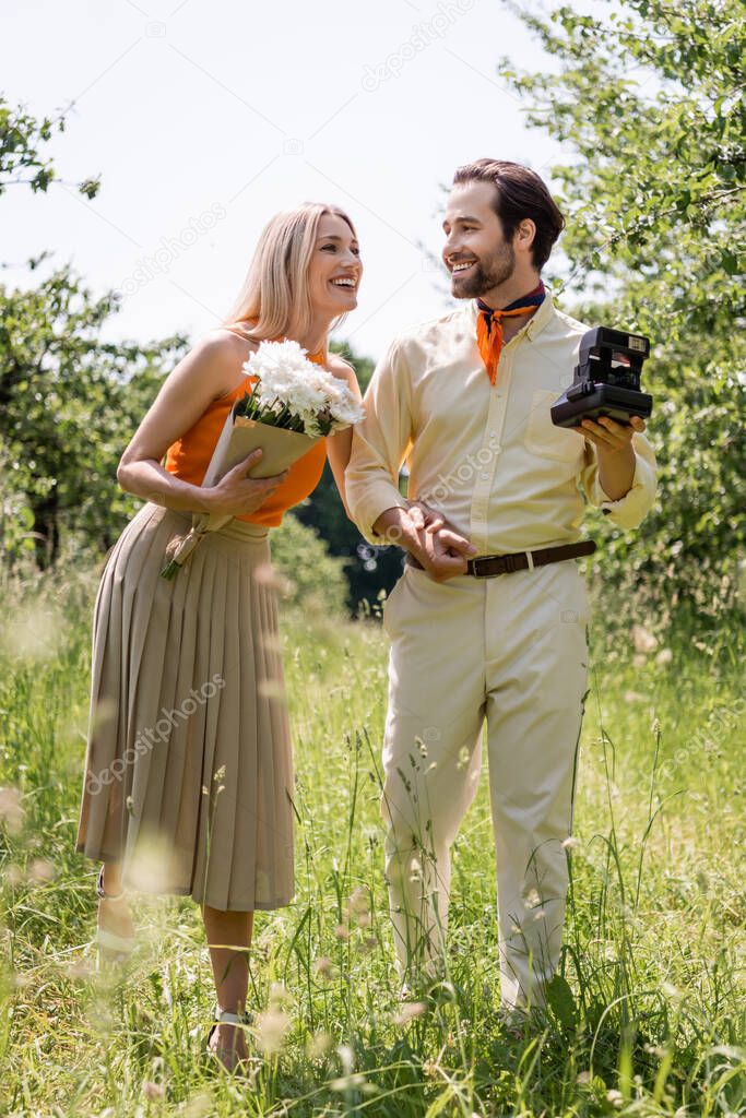 Smiling stylish couple with bouquet and vintage camera holding hands in park 