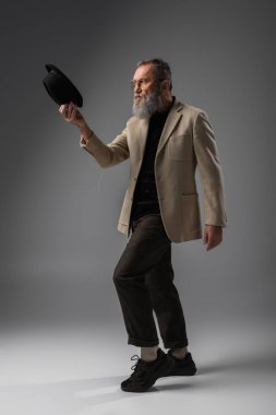 full length of bearded senior man in beige jacket and eyeglasses posing with derby hat on grey clipart