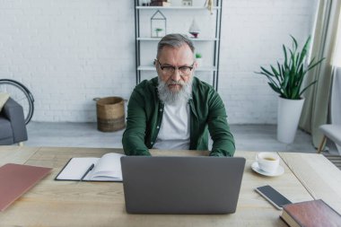 Düşünceli ve sakallı gözlüklü yaşlı adam evde çalışırken laptop kullanıyor. 