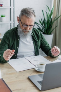 Gözlüklü sakallı yaşlı adam evden çalışırken bir fincan kahve ve dizüstü bilgisayarın yanındaki belgelere bakıyor. 