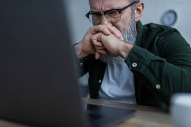 Stresli gözlüklü yaşlı adam kaşlarını çatıyor ve evdeki bulanık laptopa bakıyor.