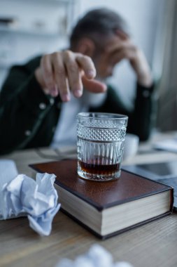blurred senior man reaching glass of whiskey on book near crumpled paper  clipart