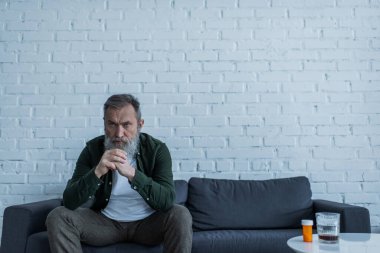 Bunalımlı, gri saçlı yaşlı bir adam. Şişenin yanındaki koltukta oturuyor. Yanında ilaç ve sehpada bir bardak viski var.