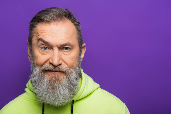 skeptical senior man moving eyebrow while looking at camera on purple background 