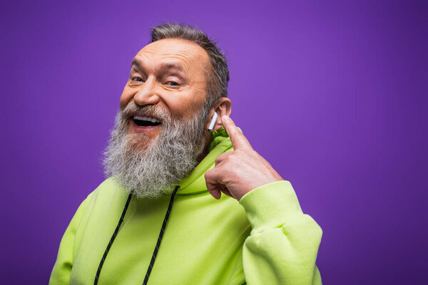cheerful senior man in green hoodie listening music and pointing with finger at wireless earphone on purple