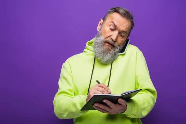 Homem Sênior Com Capuz Verde Escrevendo Notebook Falando Smartphone Roxo — Fotografia de Stock