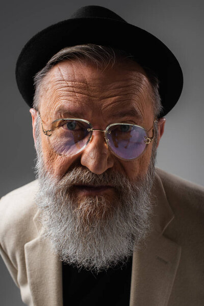 портрет стильного мужчины в очках и шляпе дерби, позирующего на сером