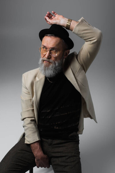 бородатый мужчина в шапке дерби и очках сидит и смотрит на серый