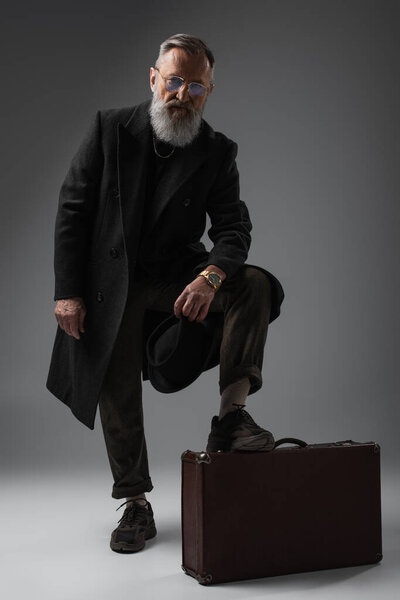 полная длина элегантного пожилого человека в стильном пальто дерби шляпу и положить ногу на чемодан на серый