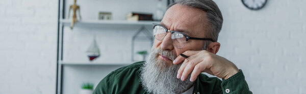 pensive and bearded senior man in eyeglasses holding pen and looking away at home, banner