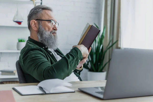 Bärtiger Senior Mit Brille Liest Buch Neben Laptop Auf Schreibtisch — Stockfoto