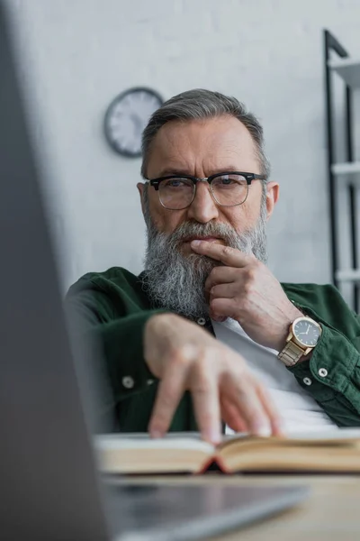 戴眼镜的体贴入微的大胡子老人一边看着笔记本电脑 一边用手触摸书本 — 图库照片