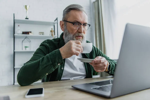 Hombre Mayor Barbudo Anteojos Bebiendo Café Mirando Ordenador Portátil Mientras — Foto de Stock