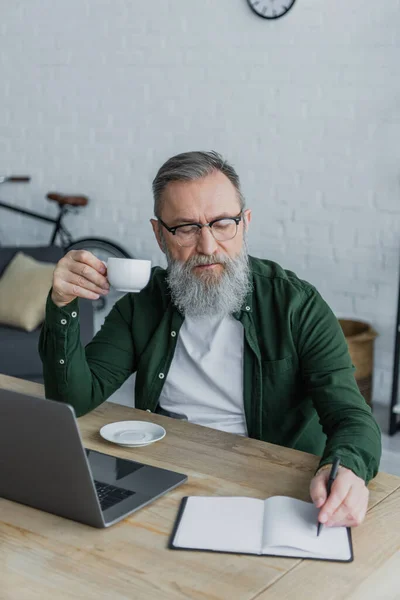 Bebaarde Senior Man Bril Met Kopje Koffie Schrijven Buurt Laptop — Stockfoto