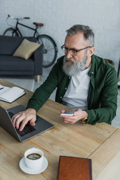 Homem Sênior Barbudo Óculos Segurando Smartphone Enquanto Usa Laptop Casa — Fotografia de Stock