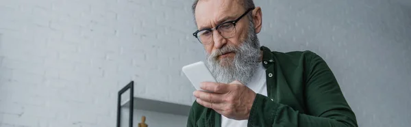 戴眼镜 留着胡子的老人在家里用智能手机 打着横幅 — 图库照片