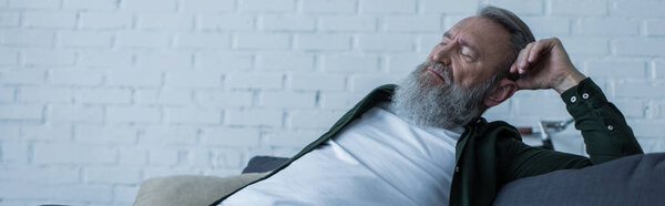 расстроен старший мужчина с бородой страдает кризисом, сидя на диване в гостиной, баннер 