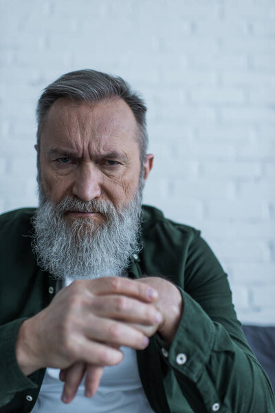 displeased senior man with beard frowning while suffering crisis and looking at camera 