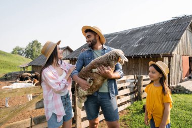 happy woman looking at husband standing with lamb on cattle farm clipart