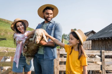 woman and child in straw hats stroking lamb in hands of smiling farmer  clipart
