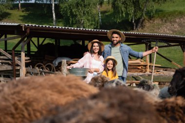 Hasır şapkalı aile bulanık ön planda ağıl ve çiftlik hayvanlarının yanında gülümsüyor