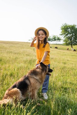 Hasır şapkalı çocuk kameraya gülümsüyor köpek ve otlakta otlayan sürü yanında.