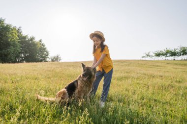 Hasır şapkalı kız yaz günü otlakta pofuduk sığır köpeğini okşuyor.