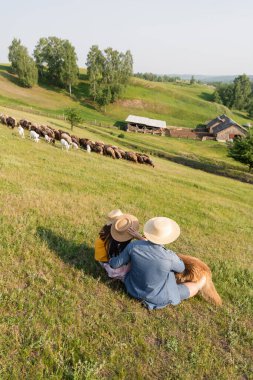 back view of family in straw hats sitting near herd grazing on scenic pasture clipart