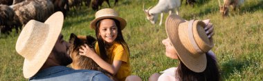 Hasır şapkalı kız sığır köpeğinin yanında gülümsüyor ve ailesi yeşil çayırda, pankartta