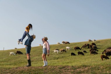side view of farmer raising up daughter near wife and herd grazing in green pasture clipart
