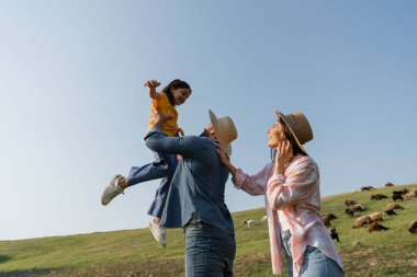 Çiftçinin yanında gülümseyen bir kadın mavi gökyüzünün altındaki yeşil çayırda çocukla oynuyor.