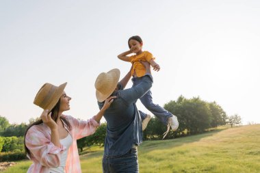 man in straw hat raising up cheerful daughter near happy wife  clipart