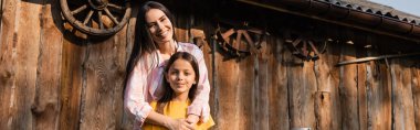 cheerful woman embracing daughter near wooden barn on farm, banner clipart