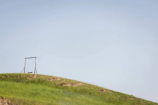 wooden swing on grassy hill under cloudless sky on summer day