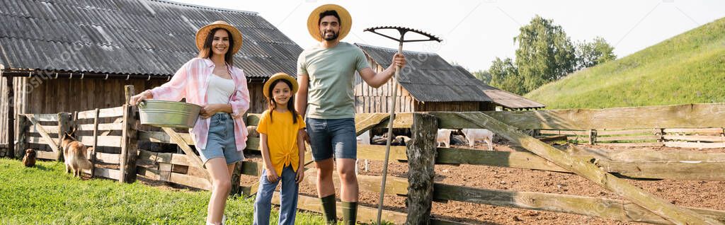 family with bowl and rakes smiling at camera near corral on rural farm, banner