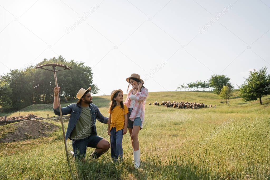 farmer holding rakes near smiling family and herd grazing in pasture