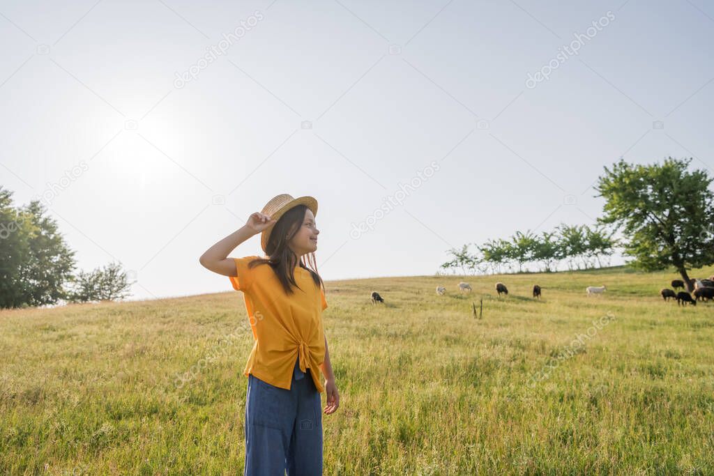 happy child adjusting straw hat and looking away in scenic meadow near grazing flock